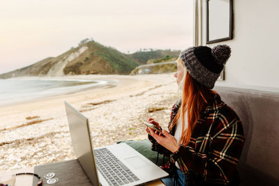 High angle of female traveling freelancer sitting in parked truck with smartphone and laptop and working on remote project while looking away