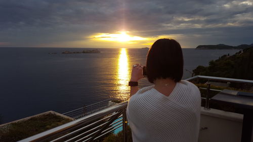 Woman standing on railing by sea against sky during sunset