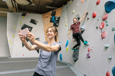 Smiling female taking selfie through smart phone with male friend climbing wall in gym