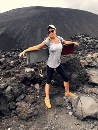 Portrait of young woman carrying metal while standing on volcanic landscape against sky