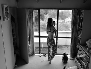 Full length rear view of woman with cats looking through window during monsoon