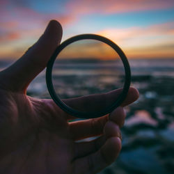 Cropped image of hand holding lens at beach during sunset