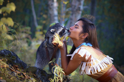 Side view of young woman in traditional clothing kissing raccoon in forest