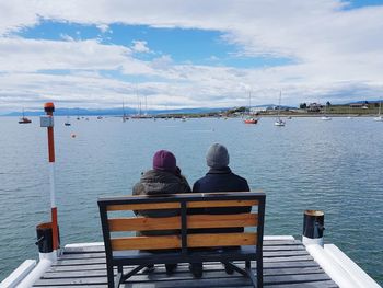 Rear view of people sitting on bench by sea against sky