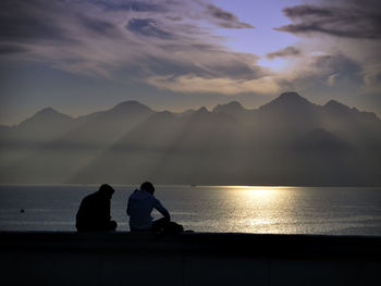 Silhouette men sitting by sea against sky during sunset