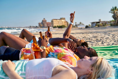 Side view of young multiethnic male and female friends in casual clothes drinking beer and pointing at cloudless blue sky while lying on sandy beach during summer holidays