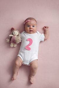 Girl baby infant with a bow lies on the bed in a white bodysuit with a doll toy next to it