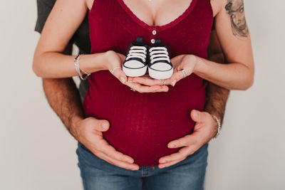 Midsection of pregnant couple holding shoes while standing against wall