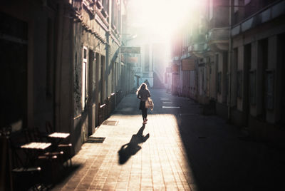 Rear view of woman walking on footpath amidst buildings during sunny day