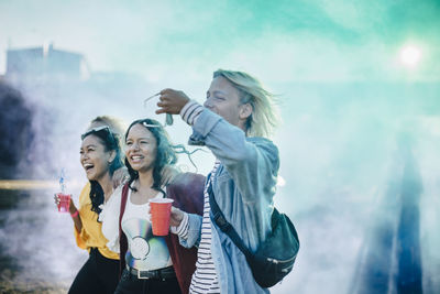 Cheerful friends enjoying drinks while walking amidst powder paint at music festival