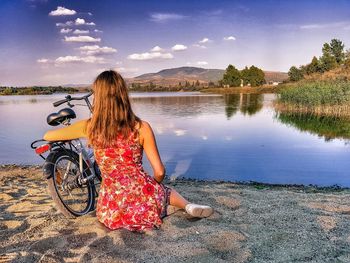 Woman sitting on bicycle by lake against sky