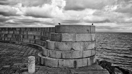 Retaining wall by sea against cloudy sky