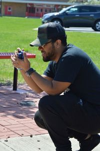 Side view of young man photographing through camera while crouching on footpath