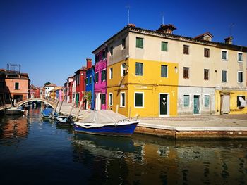 Boats moored in canal by buildings against clear blue sky