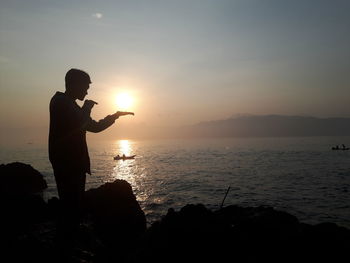 Silhouette man standing in sea against sky during sunset