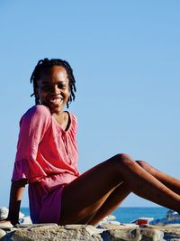 Portrait of smiling woman sitting at beach against clear blue sky