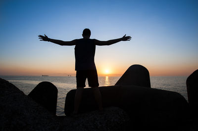 Silhouette man with arms outstretched standing on rock against sea during sunset