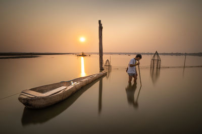 Man standing on wooden post by sea against sky during sunset