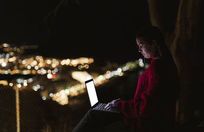 Woman sitting on hill above illuminated city at night using laptop
