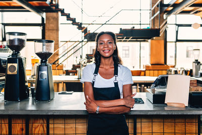 Portrait of smiling owner with arms crossed standing by bar counter in cafe