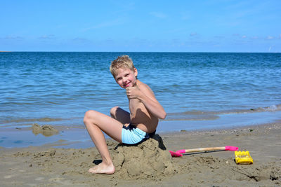 A blond boy with swimming trunks sits laughing on a pile of sand on the beach, with thumbs up