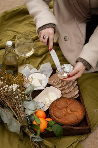 From above anonymous woman in elegant coat smearing fancy cheese on piece of bread over tray with fresh tangerines and yummy pie while sitting on blanket near wine and bouquet of flowers