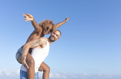 Low angle view of man piggybacking cheerful girlfriend against blue sky