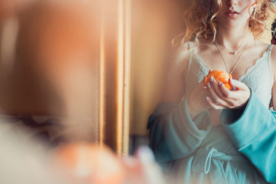 Midsection of young woman holding orange while standing by mirror at home