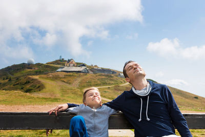 Son and father relaxing on bench against sky