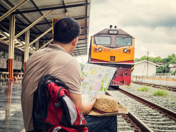 Rear view of male tourist holding map while sitting at railroad station platform