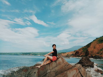 Young woman sitting on rock by sea against sky