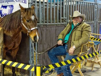 Man wearing hat sitting on seat by horse against animal pen