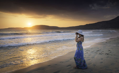 Woman photographing at sea shore against sky during sunset