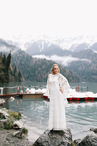 A young woman in a white lace wedding dress stands in the rain among the sea and mountains
