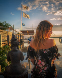 Rear view of woman sitting by buddha statue on shore against sky during sunset