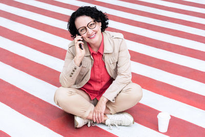 An adult woman with glasses with black hair sits on the sidewalk talking on the phone 