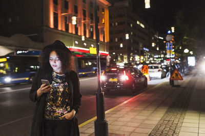 Woman using smart phone while standing on sidewalk in city at night