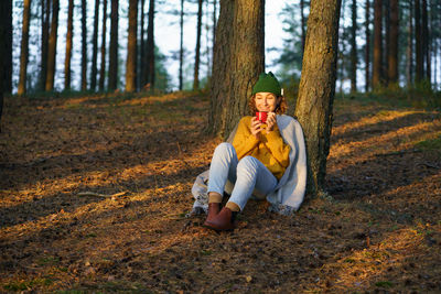 Recreation in nature. happy woman sit under tree in autumn forest drinking hot tea enjoying sunset