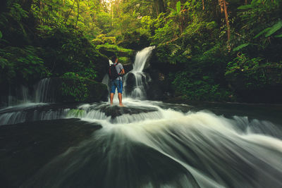 Full length of man standing on rocks against waterfall in forest