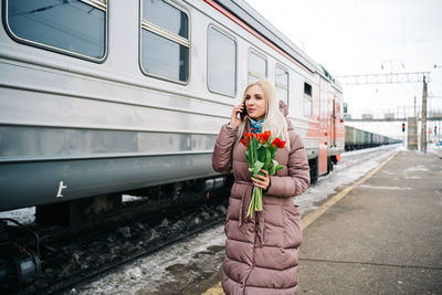 Joyful girl at the train station with flowers talking on the phone