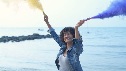 Happy young woman with arms raised on beach against sky