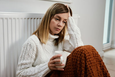 Worried woman sit near heating radiator under blanket with cup of tea