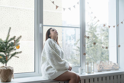 Woman in warm white winter sweater sitting on the window at home at christmas 