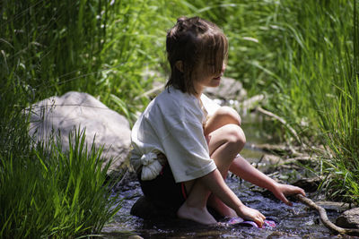 Side view of girl holding fabric while crouching amidst stream