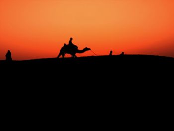 Silhouette people riding horse on shore against orange sky