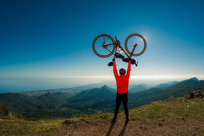 Succesful adult male cyclist rises his bicycle over his head on top of a mountain