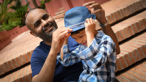Smiling father adjusting sun hat of son while sitting on steps