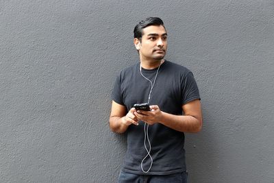 Young man looking away while listening music against wall