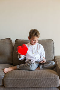Boy using mobile phone while holding heart shape sitting on sofa