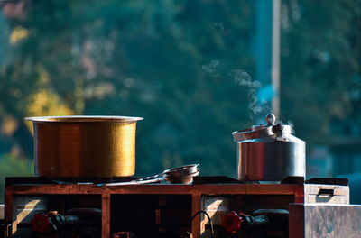 Close-up of steel teapot of chai tea on outdoor stove and golden pan of milk on green background